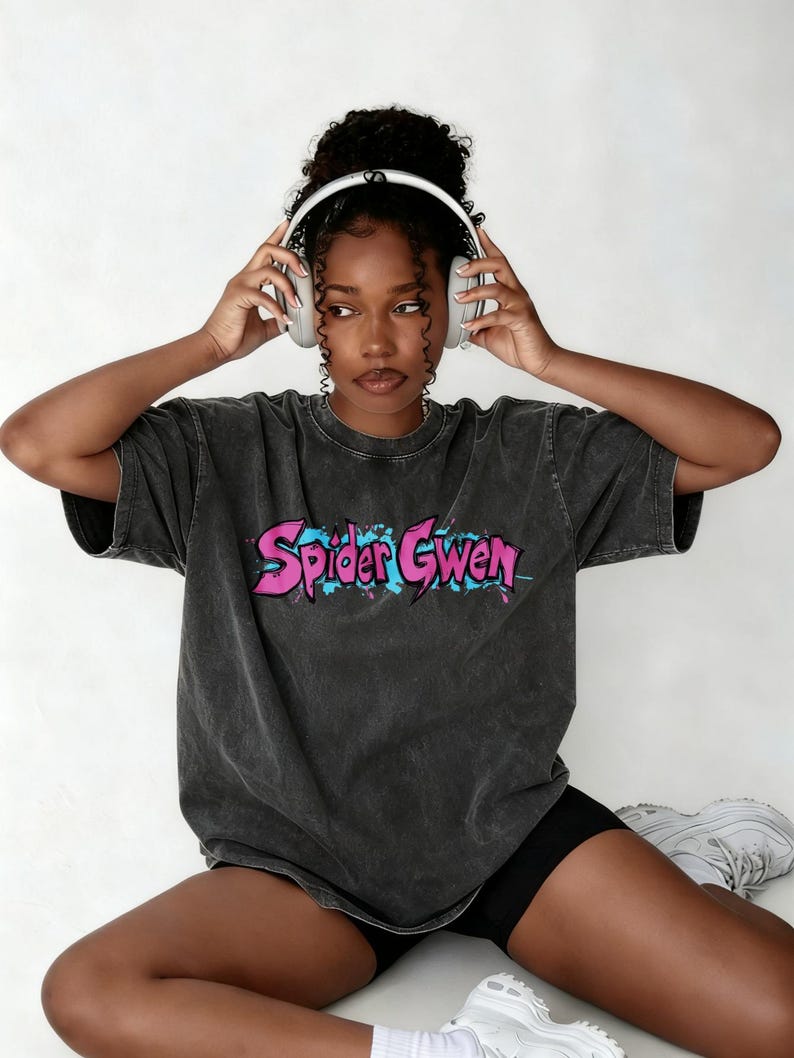 May include: A charcoal grey acid-wash t-shirt featuring the text Spider Gwen in a stylised pink font with blue paint splatter accents. The shirt has a crew neckline and short sleeves, paired with black shorts and white athletic trainers.