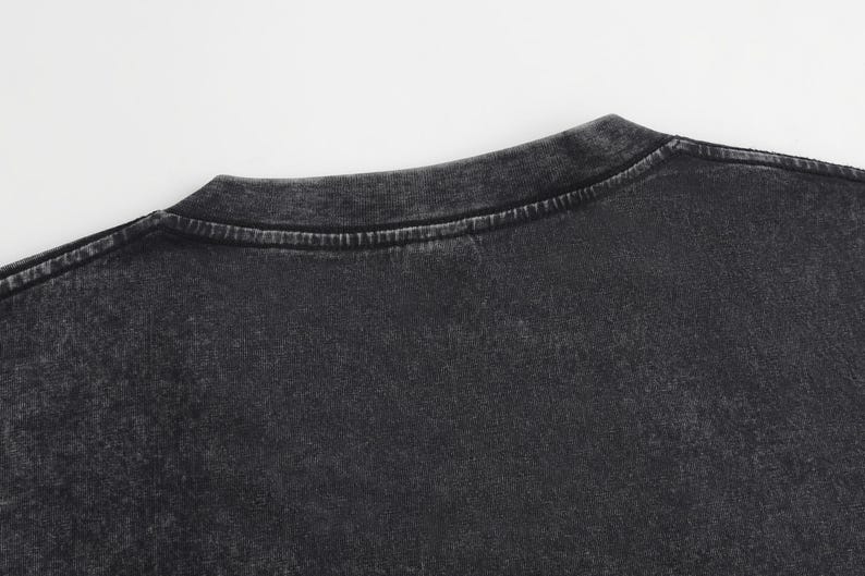 May include: A charcoal black t-shirt featuring a crew neckline and a washed-out, acid-wash texture. The fabric displays a mottled, uneven dark gray and black color pattern throughout the material, with visible stitching along the collar seam.
