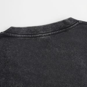May include: A charcoal black t-shirt featuring a distinct acid-wash effect that creates a mottled, vintage-inspired appearance. The garment has a classic crew neckline with a reinforced stitched collar and short sleeves, showcasing a textured finish.