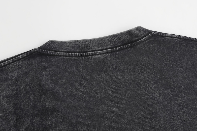 May include: A charcoal black t-shirt featuring a distinct acid-wash effect that creates a mottled, vintage-inspired appearance. The garment has a classic crew neckline with a reinforced stitched collar and short sleeves, showcasing a textured finish.