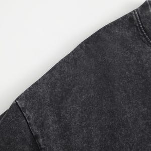 May include: A charcoal black t-shirt featuring a washed-out, mottled texture across the fabric. The garment has a classic crew neckline with visible stitching along the collar and shoulder seams, showcasing a faded, worn-in aesthetic throughout.