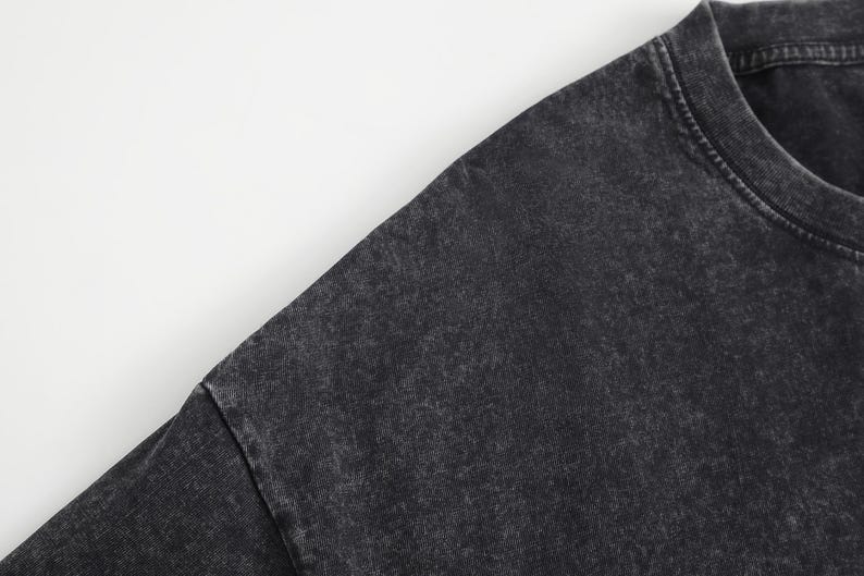 May include: A charcoal black t-shirt featuring a washed-out, mottled texture across the fabric. The garment has a classic crew neckline with visible stitching along the collar and shoulder seams, showcasing a faded, worn-in aesthetic throughout.
