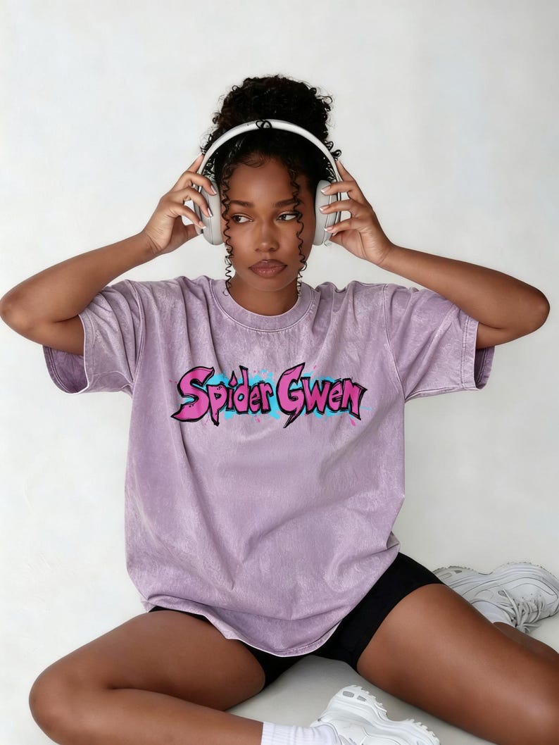 May include: A light purple mineral-washed t-shirt featuring the text Spider Gwen in a stylised pink font with a blue outline. The shirt has short sleeves and a crew neckline. The graphic is centred on the chest area of the garment.