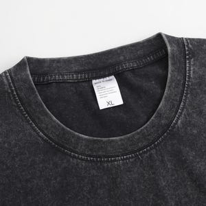 May include: A charcoal grey t-shirt with a mineral wash finish and a ribbed crew neckline. A white rectangular garment tag is attached to the inside of the collar, featuring the text MADE IN CHINA and the size XL printed in black ink.