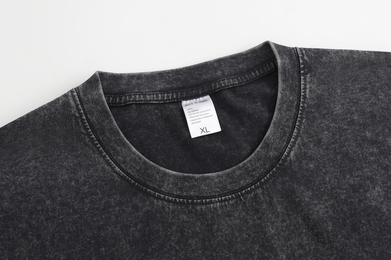 May include: A charcoal grey t-shirt with a mineral wash finish and a ribbed crew neckline. A white rectangular garment tag is attached to the inside of the collar, featuring the text MADE IN CHINA and the size XL printed in black ink.