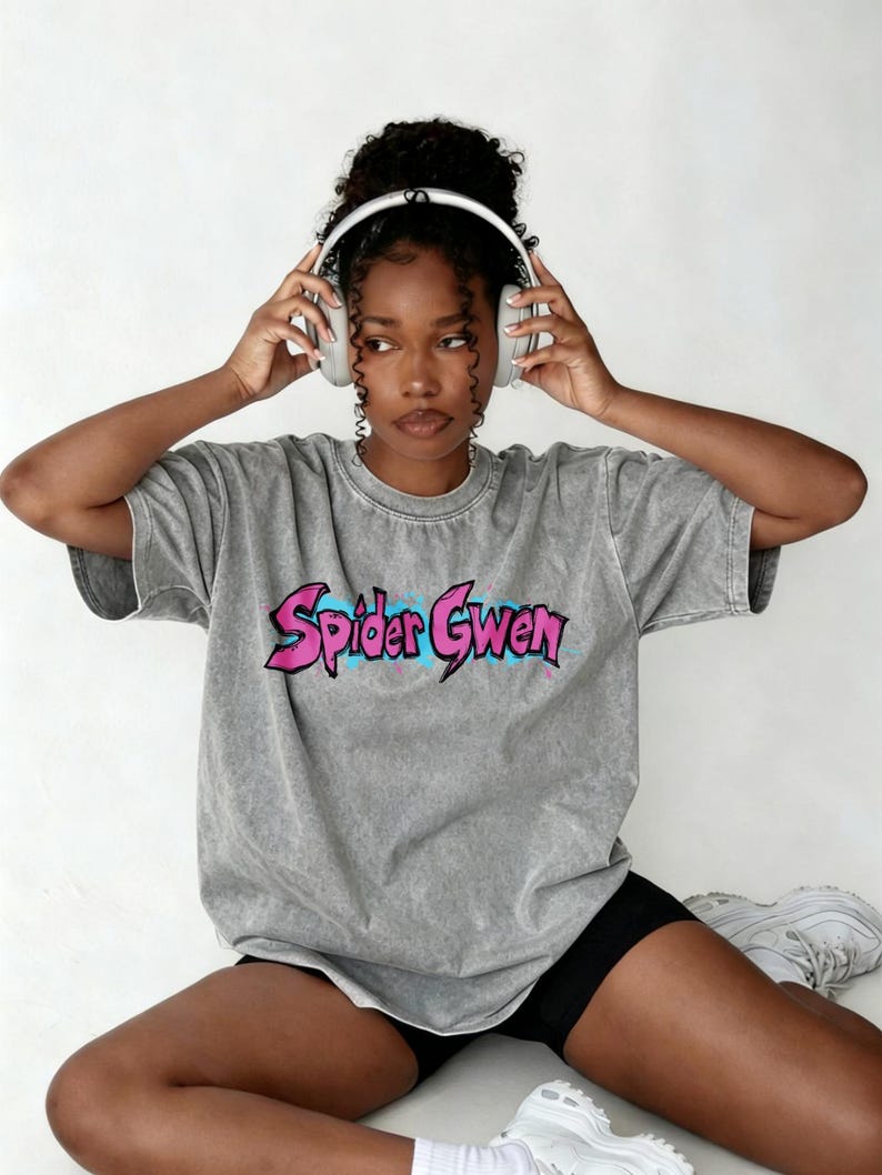 May include: A grey acid-wash t-shirt featuring the text Spider Gwen in a stylised pink font with blue paint splatter accents. The shirt has a crew neckline and short sleeves, providing a relaxed fit. The graphic is centred on the chest area.