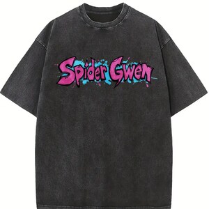 May include: A charcoal grey acid-wash t-shirt with short sleeves and a crew neckline. The chest features the text Spider Gwen in a stylised, graffiti-inspired font with bright pink lettering, blue outlines, and blue and pink paint splatters.