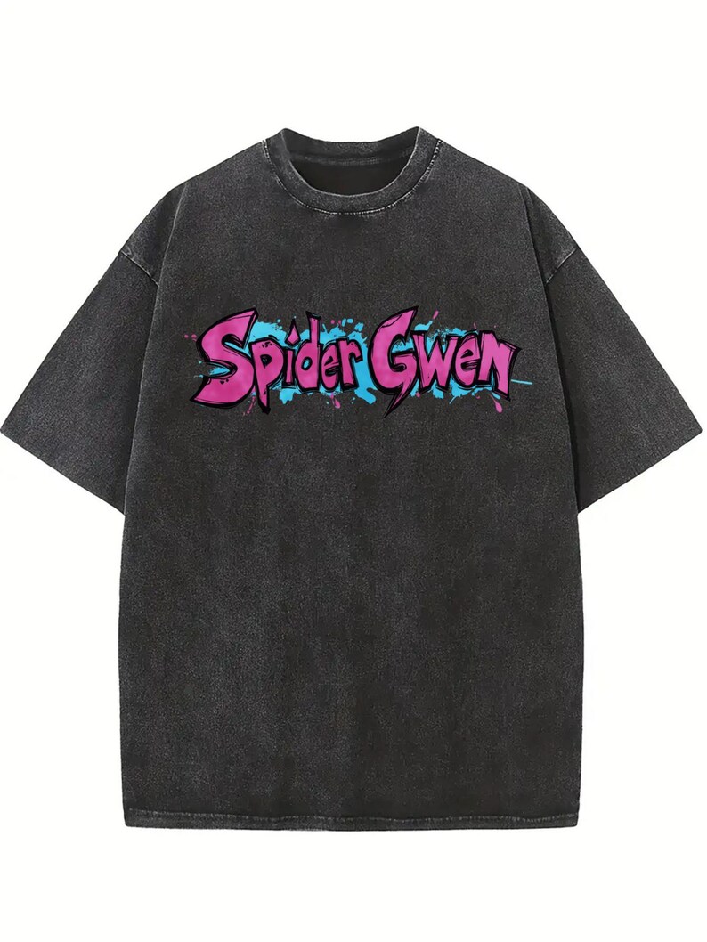 May include: A charcoal grey acid-wash t-shirt with short sleeves and a crew neckline. The chest features the text Spider Gwen in a stylised, graffiti-inspired font with bright pink lettering, blue outlines, and blue and pink paint splatters.