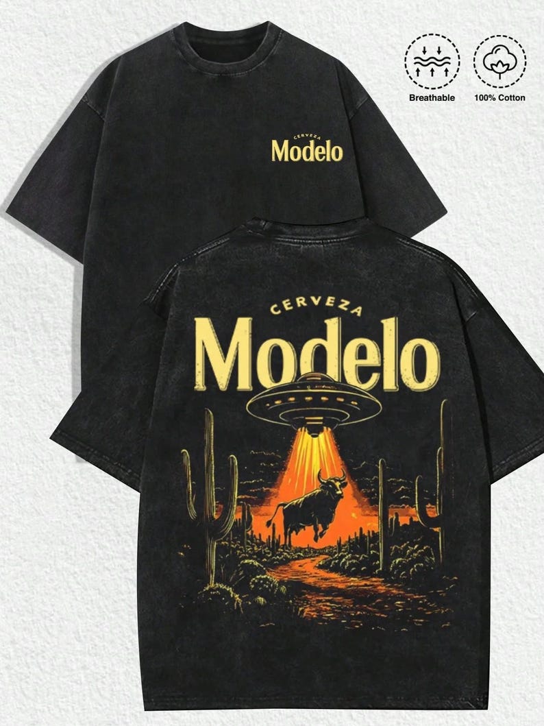 May include: A black short-sleeve t-shirt featuring a small yellow Modelo logo on the front chest. The back displays a large graphic of a bull being abducted by a UFO in a desert landscape with cacti, under the text Cerveza Modelo. Includes icons for breathable and 100% cotton.