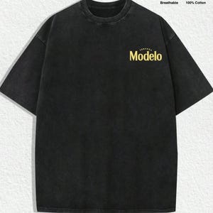 May include: A black short-sleeve t-shirt with a crew neckline and a washed-out finish. The chest features the yellow text "Cerveza Modelo". Two circular icons in the top right corner indicate the shirt is breathable and made of 100% cotton.
