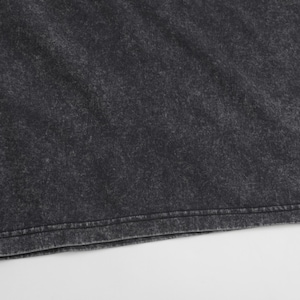 May include: A dark charcoal grey t-shirt featuring a distinct acid-wash or mineral-wash effect. The fabric displays a mottled, uneven texture with lighter grey highlights throughout the surface and a straight, double-stitched hem at the bottom edge.