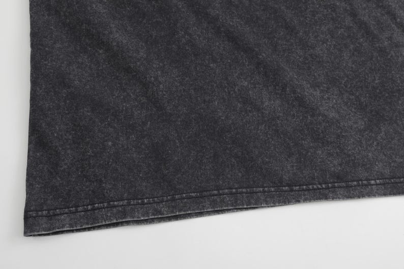 May include: A dark charcoal grey t-shirt featuring a distinct acid-wash or mineral-wash effect. The fabric displays a mottled, uneven texture with lighter grey highlights throughout the surface and a straight, double-stitched hem at the bottom edge.