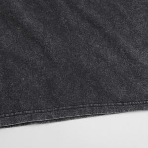 May include: A charcoal gray t-shirt featuring a mineral wash finish with a mottled, uneven texture. The fabric displays a dark, faded aesthetic with lighter gray highlights throughout the material and a double-stitched hem along the bottom edge.