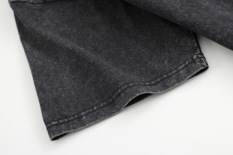 May include: A short sleeve of a garment featuring a dark charcoal grey fabric with a mottled, acid-washed texture. The sleeve has a double-stitched hem along the bottom edge, showcasing a consistent, weathered appearance across the material surface.