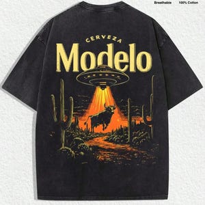 May include: A black short-sleeve t-shirt featuring a graphic print of a UFO beaming light onto a bull in a desert landscape with cacti. The text "CERVEZA" and "Modelo" appear above the UFO. Icons indicate the shirt is breathable and 100% cotton.