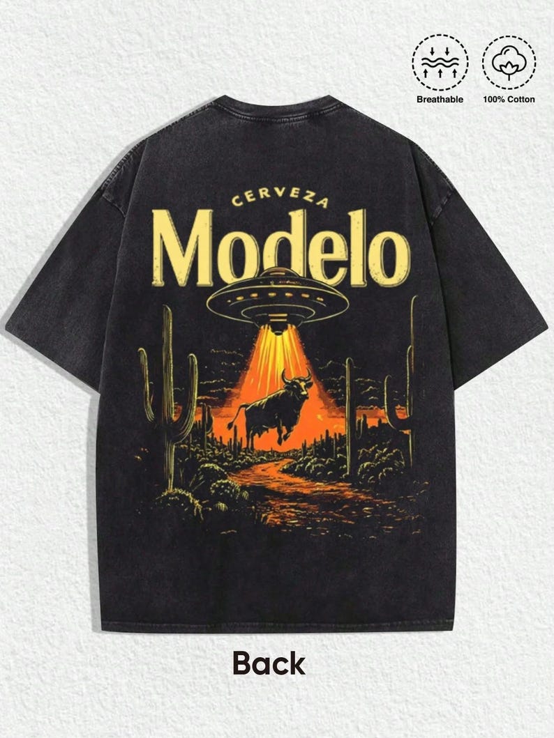 May include: A black short-sleeve t-shirt featuring a graphic print of a UFO beaming light onto a bull in a desert landscape with cacti. The text "CERVEZA" and "Modelo" appear above the UFO. Icons indicate the shirt is breathable and 100% cotton.