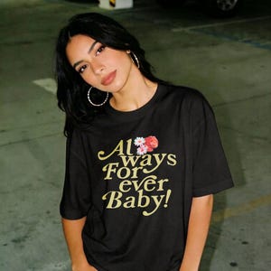 May include: A black short-sleeved t-shirt featuring the gold text Always For ever Baby! printed on the front. Above the text, there is a small graphic of a red rose and white daisies. The shirt has a crew neckline and a relaxed fit.