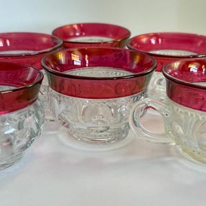 May include: A set of five clear glass cups featuring a deep red rim and a textured pattern of raised circular dots around the exterior. Each cup has a short, sturdy handle and a wide opening, showcasing a clear base and a vibrant red upper band.