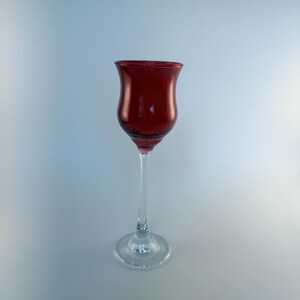 May include: A tall, slender glass with a deep red, tulip-shaped bowl and a clear, transparent stem and circular base. The vessel features a smooth, curved silhouette that tapers from the wide rim down to the narrow connection point at the stem.
