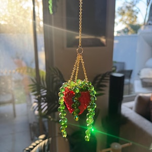 Könnte beinhalten: Ein hängendes Ornament mit einem kleinen roten, erdbeerförmigen Pflanzgefäß, das an goldfarbenen Metallketten befestigt ist. Das Gefäß enthält herabhängende Stränge aus facettierten grünen Perlen, die eine Rankenpflanze imitieren.