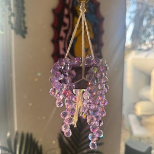 May include: A hanging ornament featuring a dark-coloured miniature pot suspended by light-coloured string. The pot is filled with cascading strands of iridescent, light purple, faceted beads that mimic the appearance of a trailing plant.