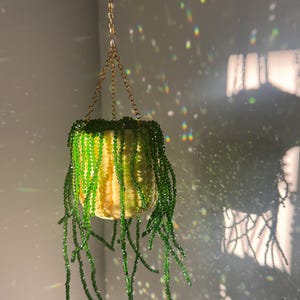 May include: A hanging planter featuring a yellow, textured pot suspended by a gold-toned metal chain. The pot contains a trailing plant made of numerous strands of small, translucent green beads that drape downwards, creating a beaded foliage effect.