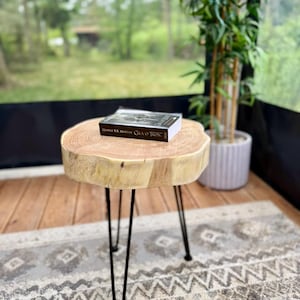 Op de afbeelding: Een ronde bijzettafel met een blad van een natuurlijke lichtgekleurde houten schijf met zichtbare nerven en schorsranden, ondersteund door drie dunne zwarte metalen haarspeldpoten. Een boek getiteld A Game of Thrones ligt op het houten blad.