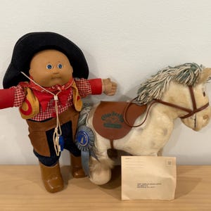 Könnte beinhalten: Eine Cabbage Patch Kids Puppe als Cowboy mit schwarzem Hut, rot-weiß kariertem Hemd und brauner Weste, neben einem weißen Plüschpferd mit braunem Sattel, einer blauen Schleife und einem kleinen Umschlag mit aufgedrucktem Text.
