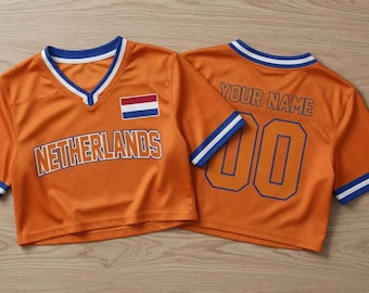 May include: Two orange cropped jerseys with blue and white trim. The front features the text NETHERLANDS and a Dutch flag. The back displays the text YOUR NAME and the number 00 in blue outline. The jerseys have V-neck collars and short sleeves.