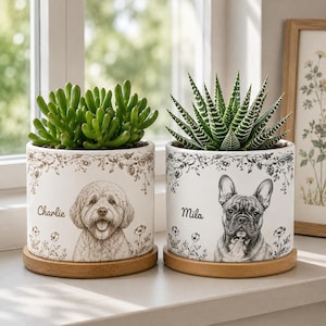 May include: Two white ceramic cylindrical planters with wooden saucers. The left planter features a brown illustration of a dog with the name Charlie. The right planter features a black illustration of a French Bulldog with the name Mila.