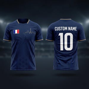 K&ouml;nnte beinhalten: Marineblaues Fu&szlig;balltrikot mit goldenen Verzierungen, das die franz&ouml;sische Flagge und die Umrisse des Eiffelturms zeigt. Die Vorderseite zeigt ein dezentes Muster und die Nummer 10 auf dem R&uuml;cken, mit "CUSTOM NAME" dar&uuml;ber. Ein stilvolles Design f&uuml;r Sportbegeisterte.