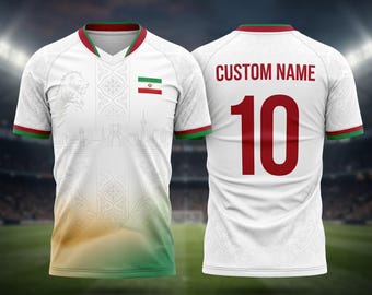 May include: White soccer jersey with green and red trim, featuring the Iranian flag and a lion graphic. The back displays "CUSTOM NAME" above the number 10 in red. The jersey has a V-neck design and a subtle pattern.