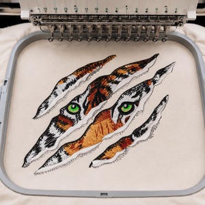 Könnte beinhalten: Ein Stickdesign auf cremefarbenem Stoff, das in einem quadratischen Rahmen eingespannt ist und ein Tigergesicht zeigt, das durch drei diagonale Krallenspuren sichtbar wird. Der Tiger hat orangefarbenes, schwarzes und weißes Fell sowie zwei leuchtend grüne Augen.