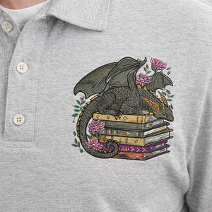 Könnte beinhalten: Ein graues meliertes Poloshirt mit einer Stickerei, die einen dunkelgrünen Drachen auf einem Stapel bunter Bücher zeigt. Der Drache ist von kleinen rosa Blüten und grünen Blättern umgeben, wobei sich das Motiv im Brustbereich des Shirts befindet.
