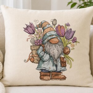 Könnte beinhalten: Ein quadratisches, cremefarbenes Zierkissen mit einer Stickerei eines Gnoms in blauem Mantel und gestreifter Mütze. Der Gnom trägt einen Terrakottatopf voller violetter Tulpen und kleiner Blumen sowie eine braune Umhängetasche an der Seite.