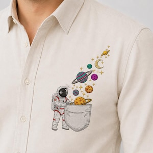 Op de afbeelding: Een beige overhemd met knoopsluiting en een borduursel van een astronaut naast een borstzak, met een spoor van kleurrijke planeten, een maansikkel en gele sterren die vanuit het zakgebied omhoog lijken te zweven.