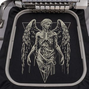 Könnte beinhalten: Ein Stickrahmen mit schwarzem Stoff und einem weißen Stickmotiv einer geflügelten Skelettfigur, die in einem Dreieck zentriert ist. Die Figur zeigt detaillierte Rippen- und Flügelstrukturen unter dem Nähfuß einer Stickmaschine.
