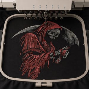 May include: An embroidery machine hoop holds black fabric featuring a stitched design of a grim reaper figure. The figure wears a red hooded robe and holds a large silver scythe. The skull is white and grey, with red thread accents.