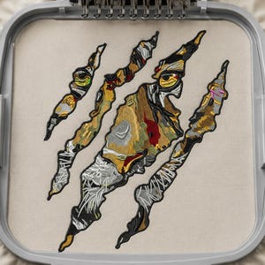 Könnte beinhalten: Ein Stickdesign mit drei gezackten Krallenspuren, die ein darunter liegendes Tigergesicht enthüllen. Das Design ist in Gelb-, Schwarz-, Weiß- und Rottönen auf beigem Stoff gestickt und wird in einem grauen Kunststoff-Stickrahmen gehalten.