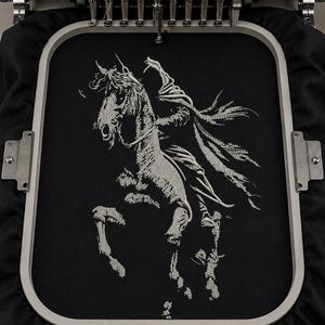 Könnte beinhalten: Ein schwarzes Stoffstück, das in einem hellgrauen Stickrahmen befestigt ist und eine detaillierte weiße Stickerei eines steigenden Pferdes mit fließender Mähne und Schweif zeigt, direkt unter den Nadeln einer Stickmaschine positioniert.