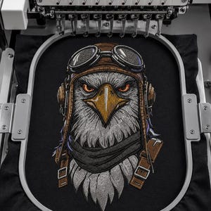 Könnte beinhalten: Ein Stickmaschinenrahmen hält einen schwarzen Stoffeinsatz mit einem detaillierten Stickmotiv eines Weißkopfseeadlers, der eine braune Fliegermütze aus Leder und eine Schutzbrille trägt. Der Adler hat einen gelben Schnabel und einen dunklen Schal.