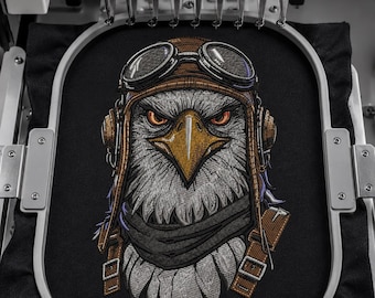 May include: An embroidery machine hoop holds a black fabric panel featuring a detailed embroidered design of a bald eagle wearing a brown leather aviator cap and goggles. The eagle has a sharp yellow beak, intense orange eyes, and a dark neck scarf.