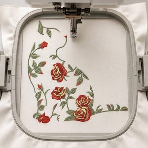 Könnte beinhalten: Ein Stickrahmen mit weißem Stoff und einem maschinengestickten Design aus roten Rosen und grünen Blättern. Das florale Muster ist in einer Eckanordnung gestaltet und zeigt mehrere blühende Rosenköpfe, Knospen und gewundene Stängel mit Laub.