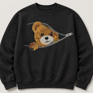 Könnte beinhalten: Ein schwarzes Langarm-Sweatshirt mit einer gestickten Grafik eines braunen Teddybären, der hinter einem weißen Reißverschluss-Design hervorschaut. Der Bär hat eine hellbraune Schnauze, schwarze Augen und eine schwarze Nase, eine Pfote ist sichtbar.