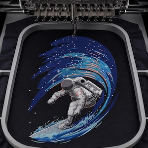 Könnte beinhalten: Ein Stickrahmen hält einen schwarzen Stoff mit einem detaillierten Stickmotiv, das einen Astronauten zeigt, der auf einer stilisierten blauen Welle surft. Der Astronaut trägt einen weißen Anzug mit grauen Schattierungen und weißen Sternen.
