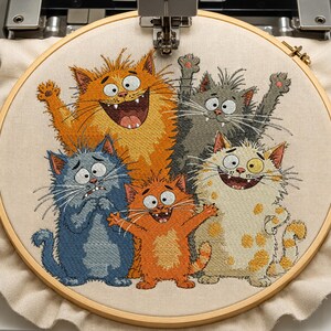 Könnte beinhalten: Ein Stickrahmen mit einem hellen Stoff, der ein gesticktes Design von fünf Cartoon-Katzen zeigt. Die Katzen sind in den Farben Orange, Grau, Blau und Creme dargestellt, mit ausdrucksstarken Gesichtern, großen Augen und offenen Mündern.