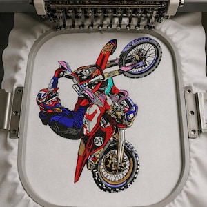 Könnte beinhalten: Ein detailliertes Maschinenstickdesign auf weißem Stoff, das einen Motocross-Fahrer zeigt, der auf einem rot-schwarzen Dirtbike einen Wheelie macht. Der Fahrer trägt einen blau-roten Helm und Ausrüstung, während das Motorrad im Stickrahmen komplexe mechanische Details und farbenfrohe Akzente aufweist.