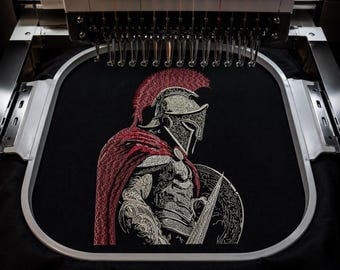 May include: An embroidery machine hoop holds a black fabric panel featuring a detailed embroidered design of a Spartan warrior. The warrior wears a silver helmet with a red crest and a red cape, holding a circular shield and a spear in profile.