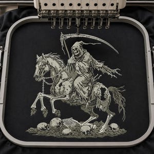 Könnte beinhalten: Ein Stickmotiv auf schwarzem Stoff, das den Sensenmann in einer Kapuzenrobe auf einem Skelettpferd zeigt. Die Figur hält eine große Sense, und die Szene umfasst einen Haufen menschlicher Schädel und Knochen an der Basis, alles in hellgrauem Garn.