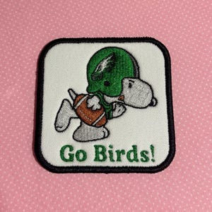 May include: A square embroidered patch with a white background and a black border. It features a cartoon dog wearing a green football helmet and holding a brown football. The text Go Birds! is embroidered in green below the cartoon character.